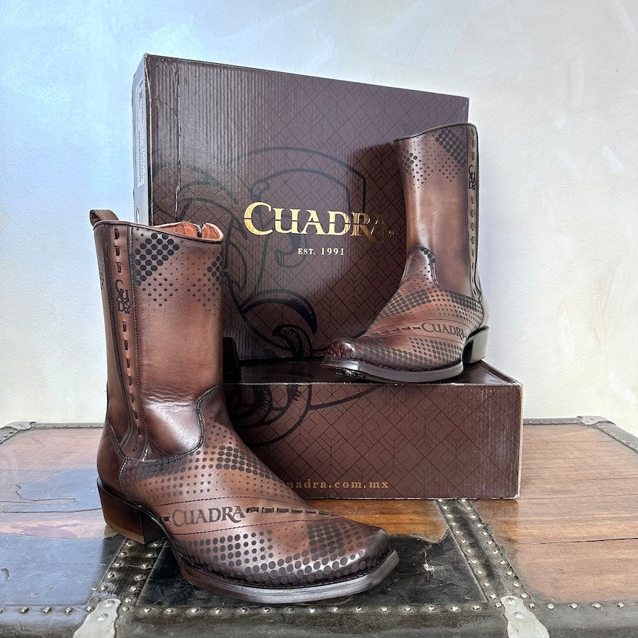 A pair of brown Cuadra men's cowhide leather ankle boots with a pointed toe and a visible brand logo on the upper, placed against a textured backdrop.