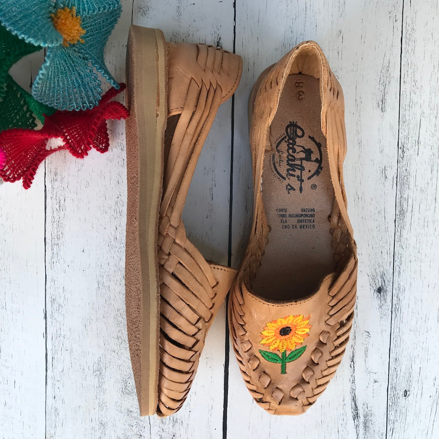 A pair of light tan, flat, leather huaraches with embroidered sunflower design on the sole, displayed on a wooden surface with a colorful decorative object in the background.